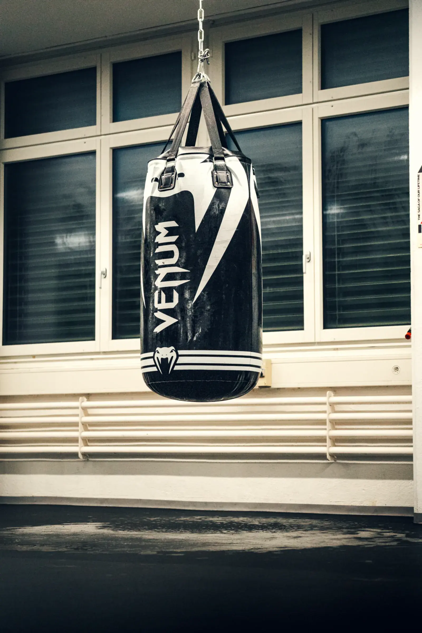 Black and white Venum heavy bag hanging on a chain in front of the gym’s windows.