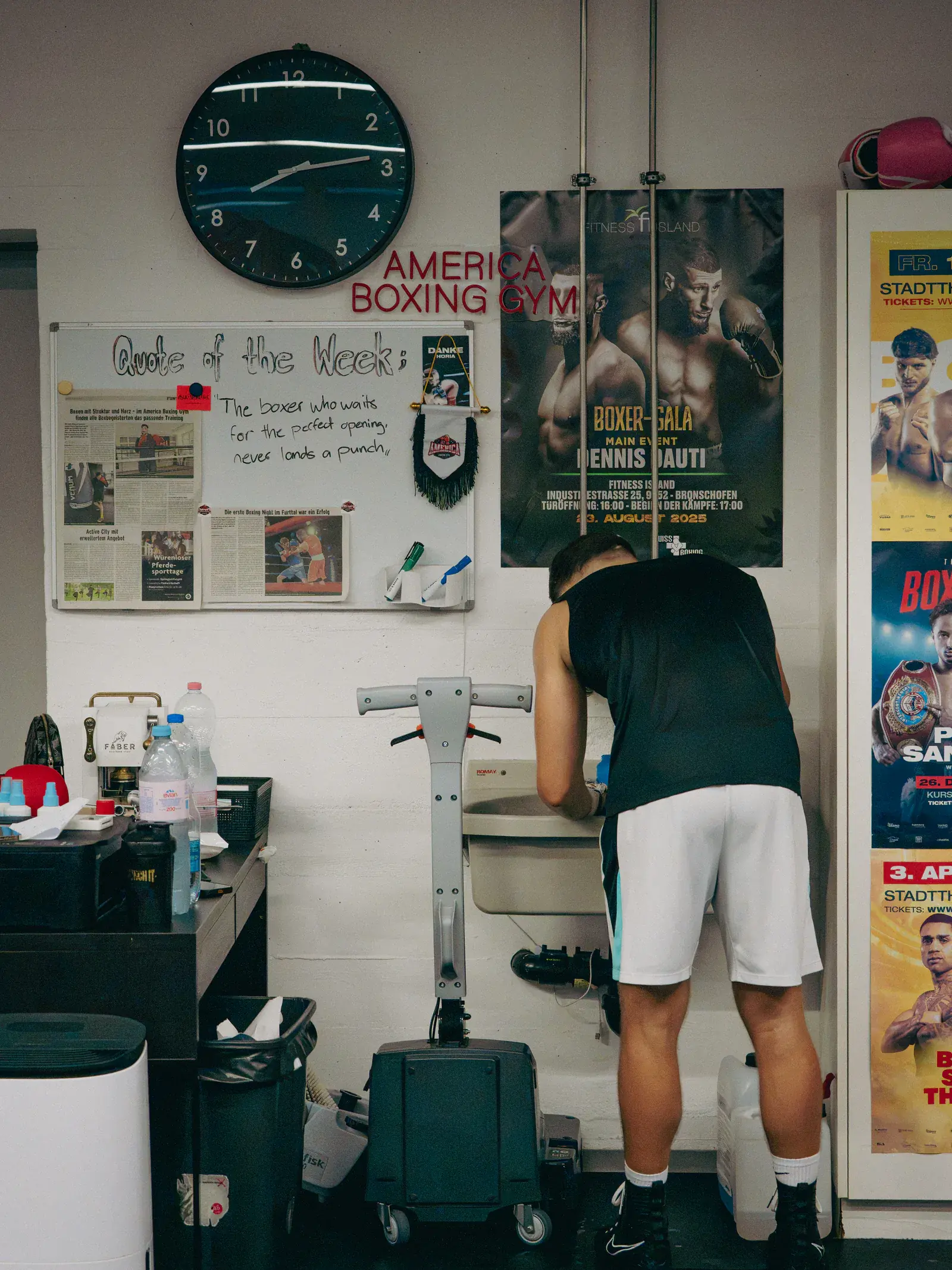 Gym wall with a large clock, the America Boxing Gym lettering, a quote-of-the-week board and boxing posters.