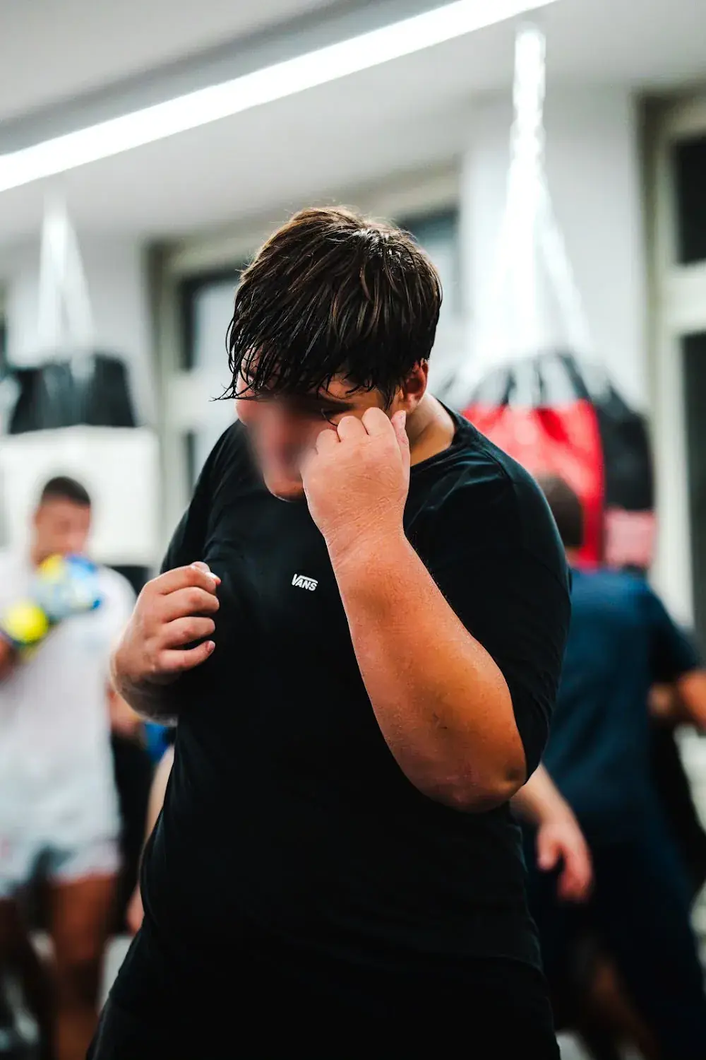 Boy in a black T-shirt with his fists raised at Kids Boxing, with heavy bags and other children behind him.