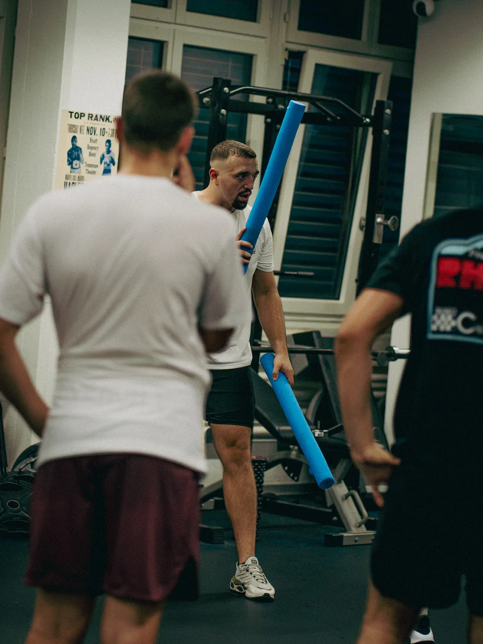 Coach in a white T-shirt holding two blue foam noodles explains a drill to boxers standing with their backs to the camera.