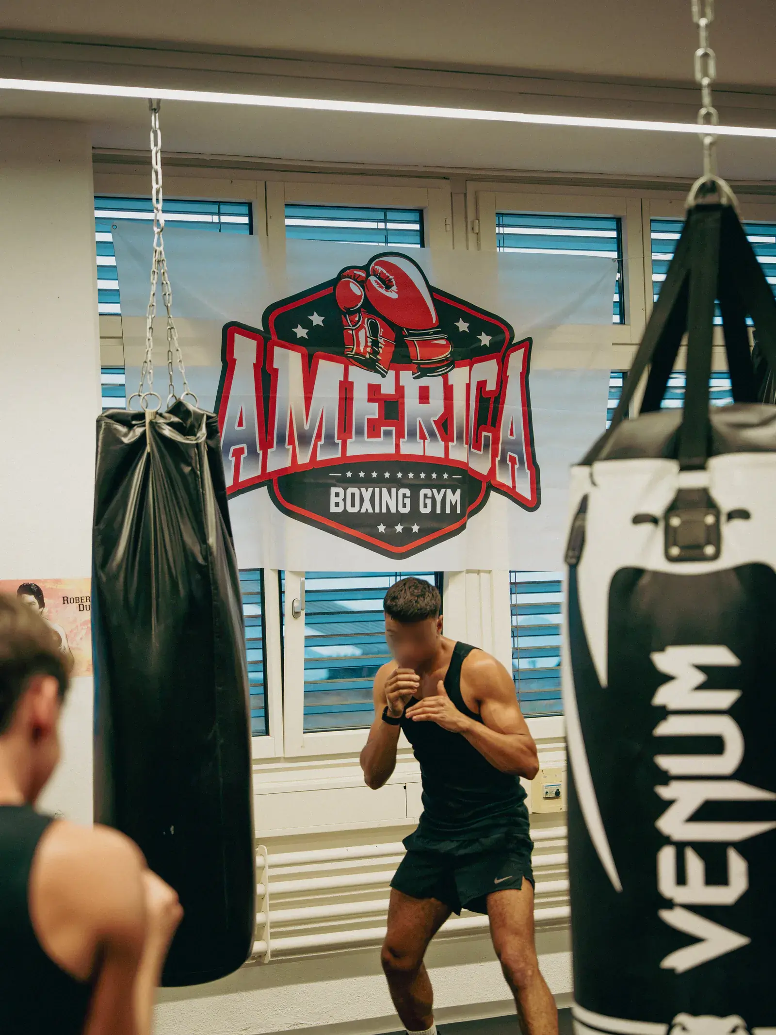 Boxer shadowboxing between two heavy bags, with the America Boxing Gym banner in the background.