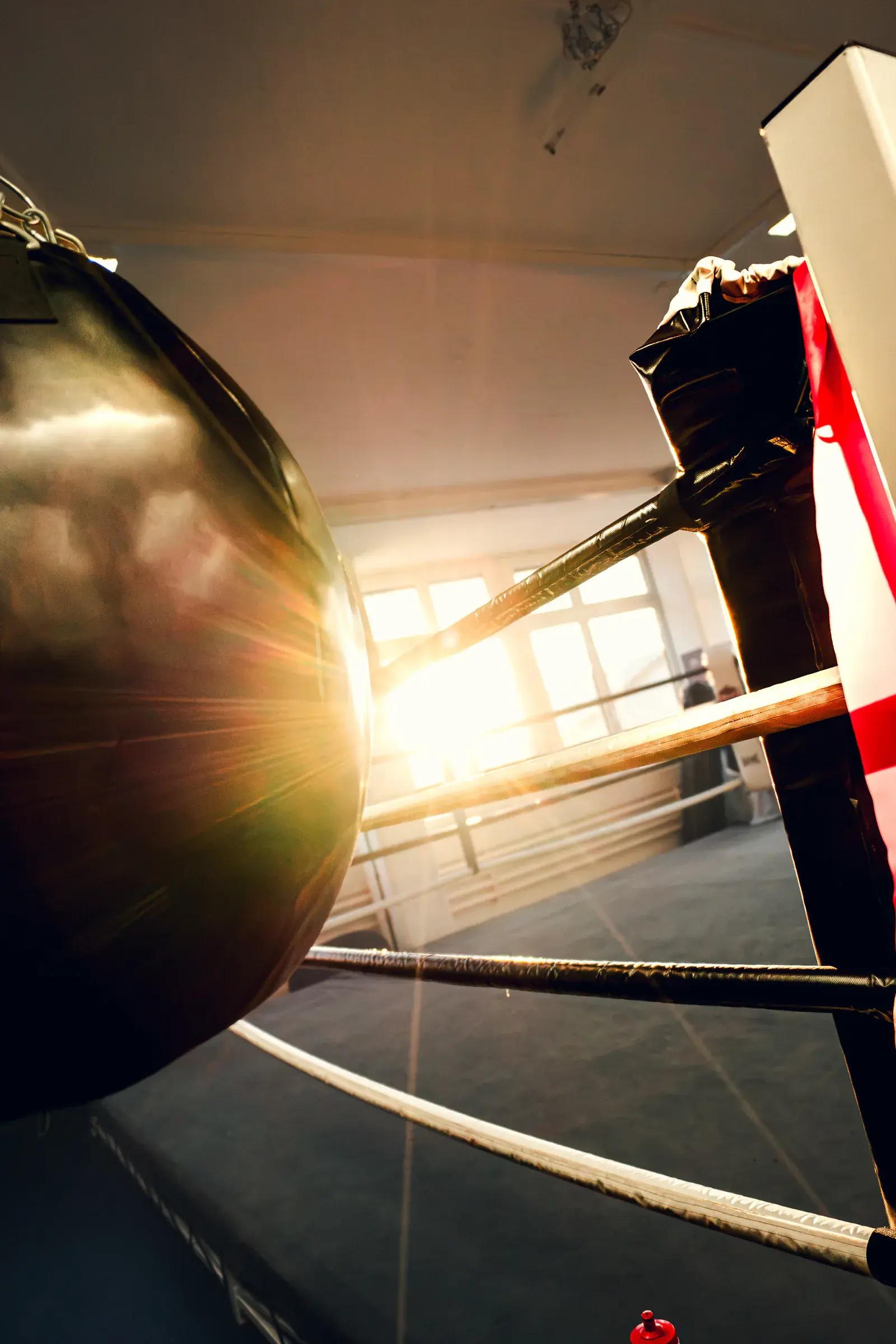 Round black bag at the corner of the ring, with the afternoon sun shining through the window onto the ropes.