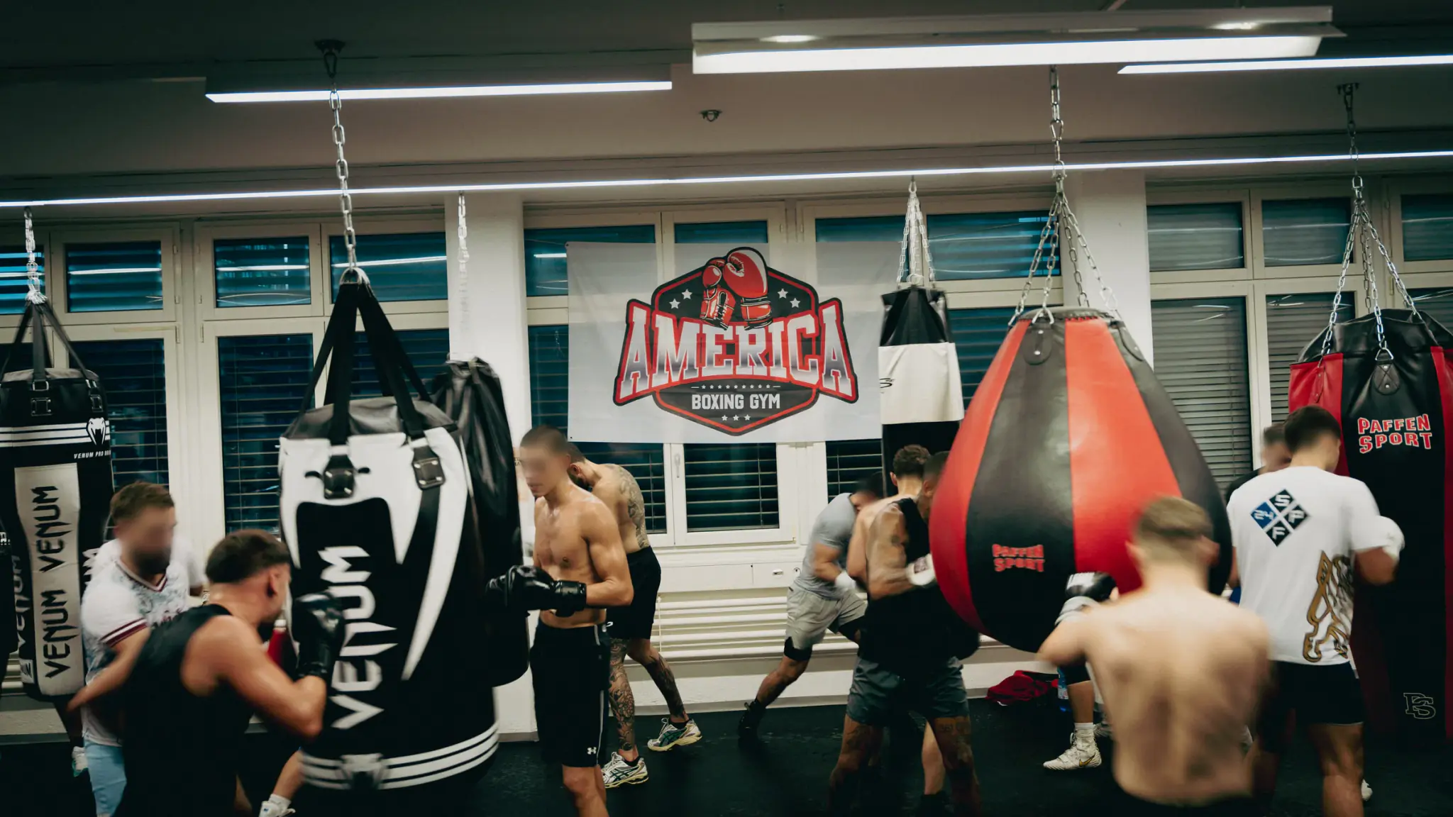 Training group at the heavy bags in America Boxing Gym, with the gym’s logo banner across the windows behind them.